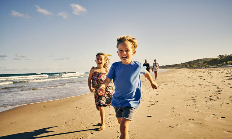 Shot of two adorable little children running at the beach with their parents in the background